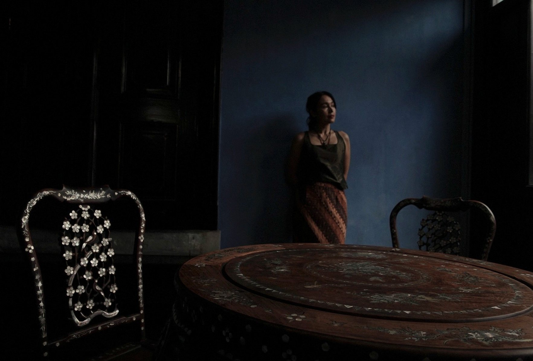 A figure standing in a darkened room with carved antique furniture, blue walls, and a single source of light.