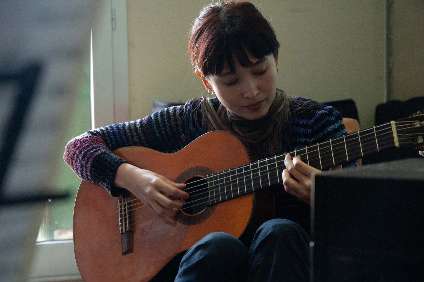 Jacelyn playing a classical guitar alone in the rehearsal space, eyes downward, concentrated, natural light from a window.