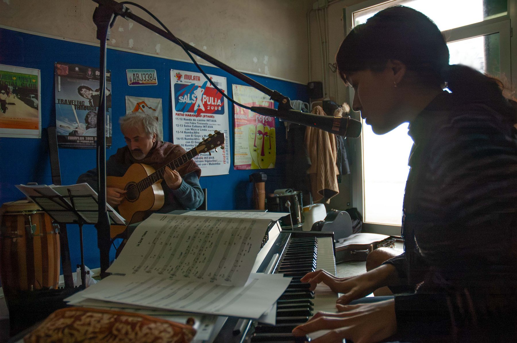 Jacelyn at a keyboard with a score open, a guitarist visible behind her in the rehearsal room, music stands and cables throughout.