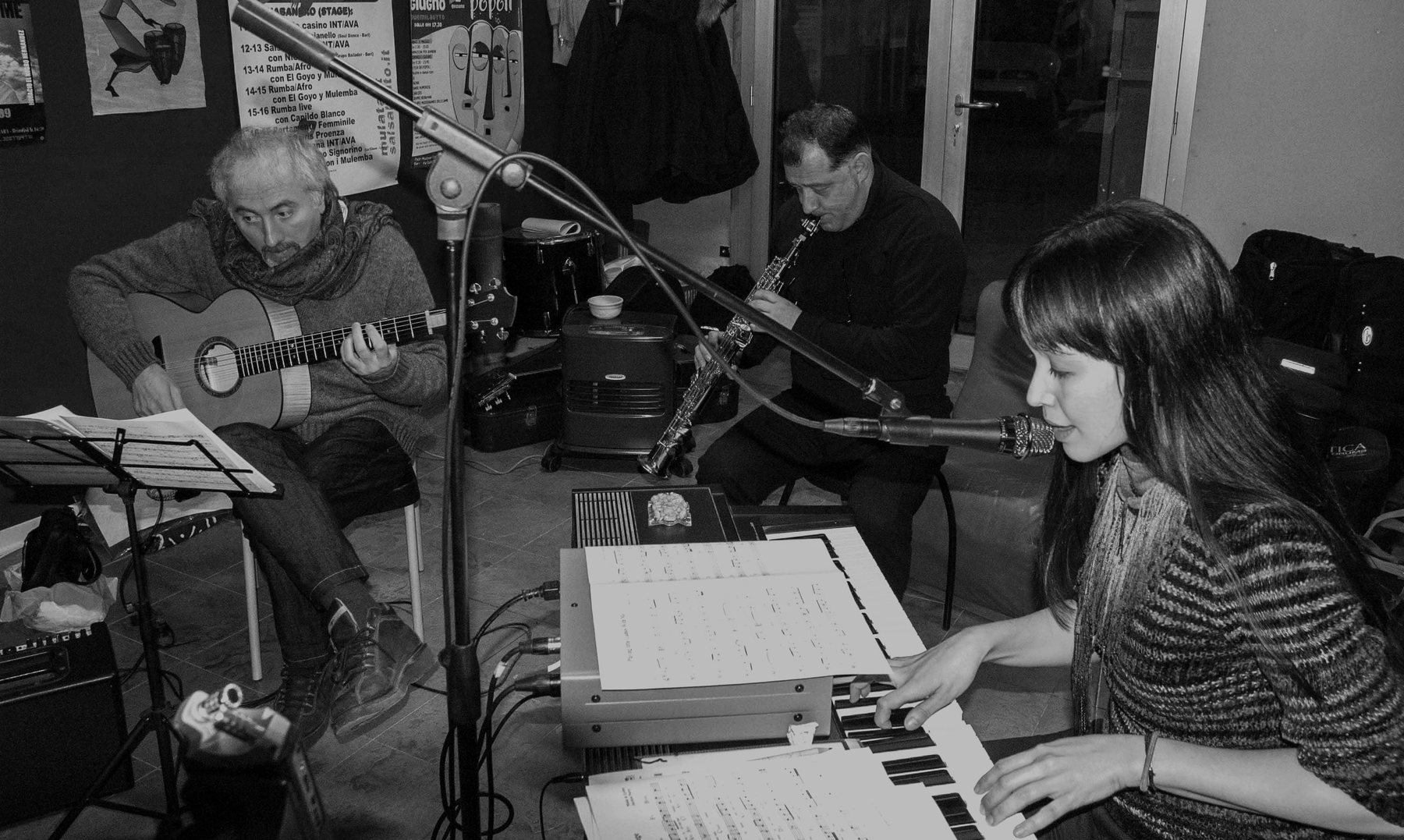 Three musicians in an evening rehearsal: guitarist to the left, soprano saxophonist in the middle, Jacelyn at keyboard on the right, scores on stands, warm light.