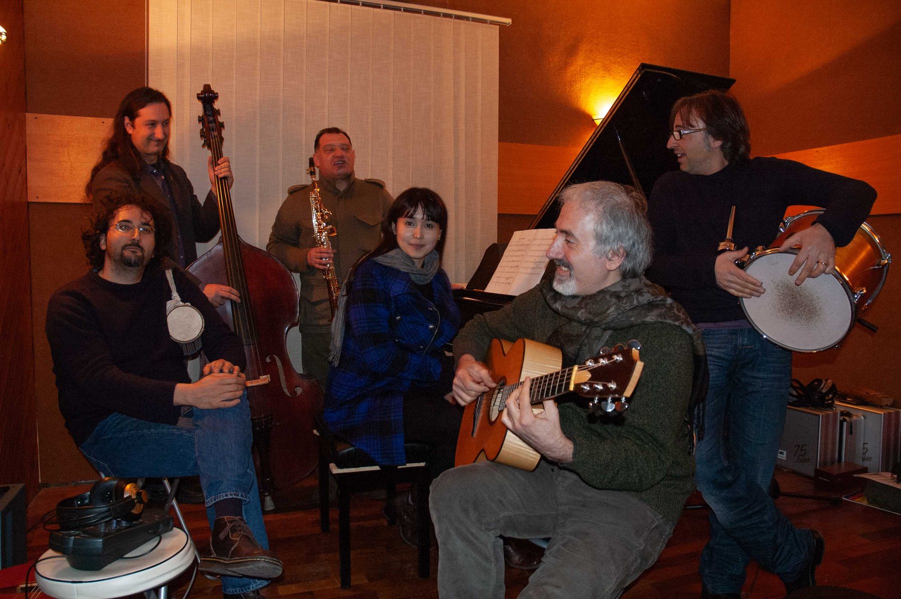 The full Fortressa ensemble in a warm wood-panelled recording studio: piano open, double bass, guitar, saxophone, percussion and tambourine, amber light throughout the room.