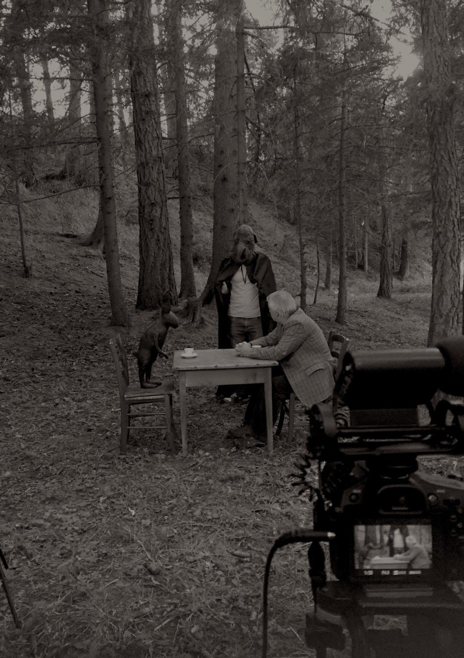 A wide view of the same forest scene, the bird-cloaked figure standing beside the seated artist, a camera monitor visible in the foreground showing the framed shot.