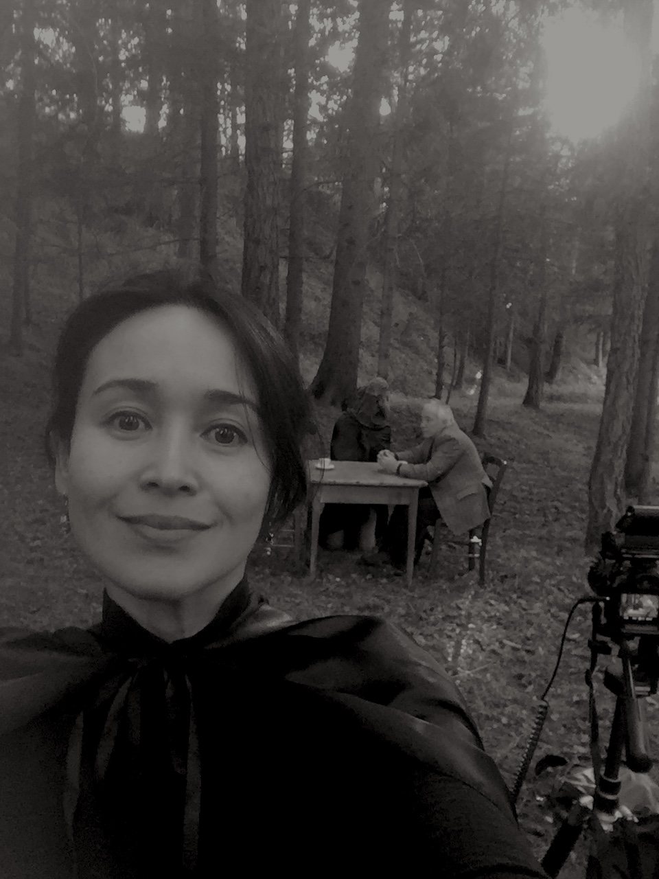 A self-portrait of the cineasti wearing the black cloak, smiling at the camera, the artist and the seated tripod visible in the forest behind her.