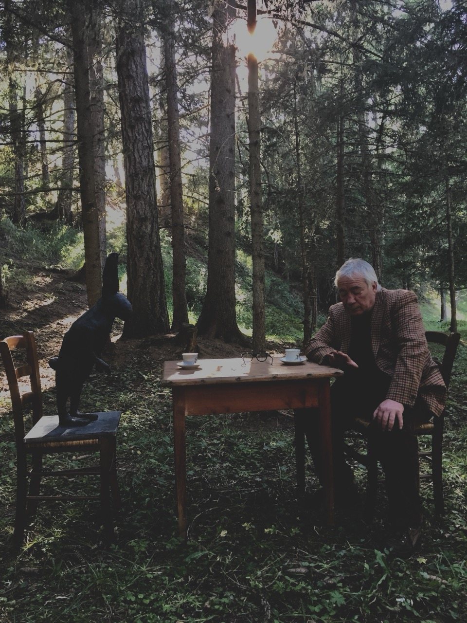 A man seated at a small wooden table set up in a forest clearing, a carved black animal sculpture on a chair beside him, sunlight through the trees.
