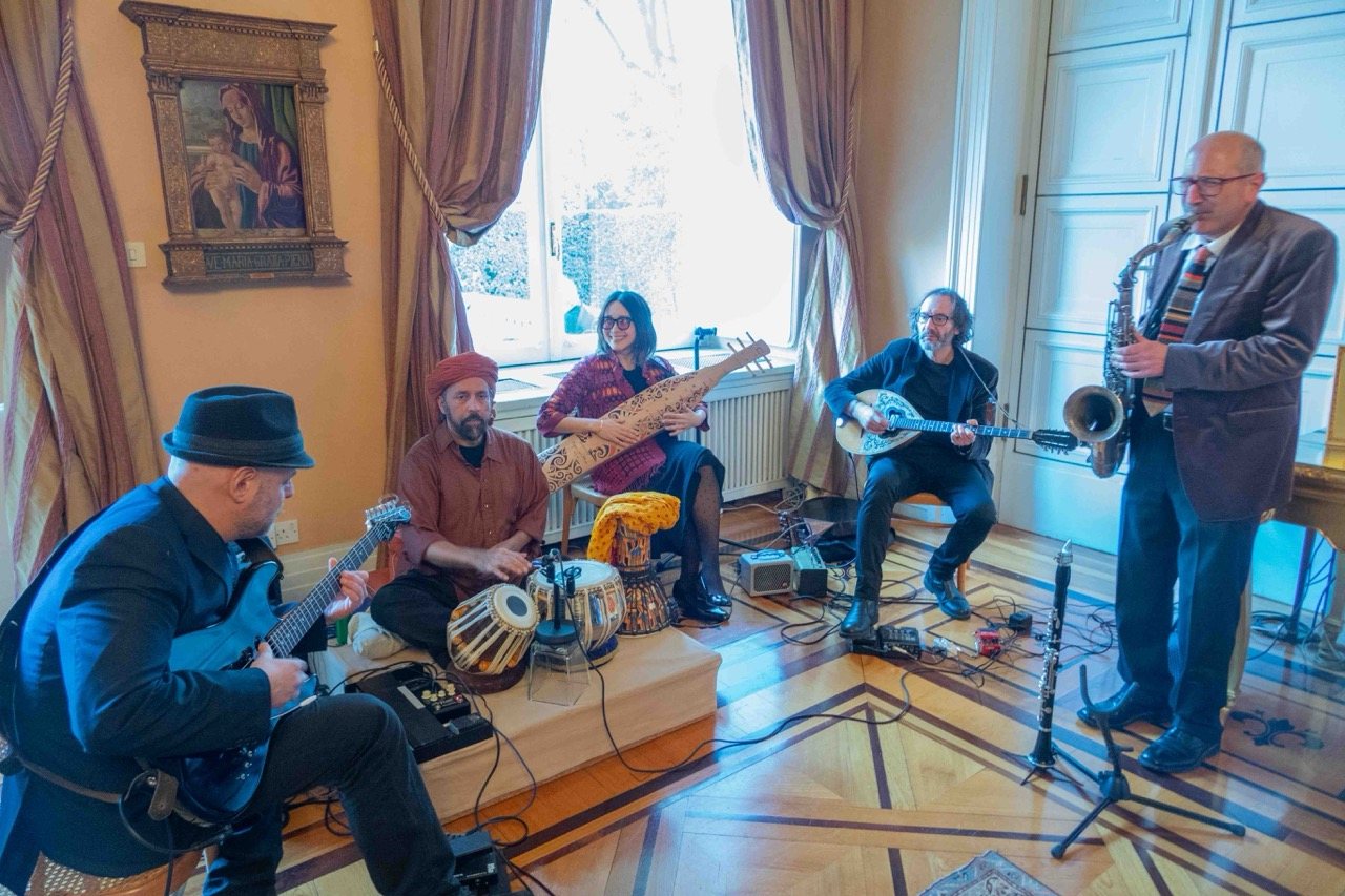 Five musicians arranged in a loose circle inside a grand room, playing guitar, tabla, sape, oud, and saxophone together beneath tall windows.