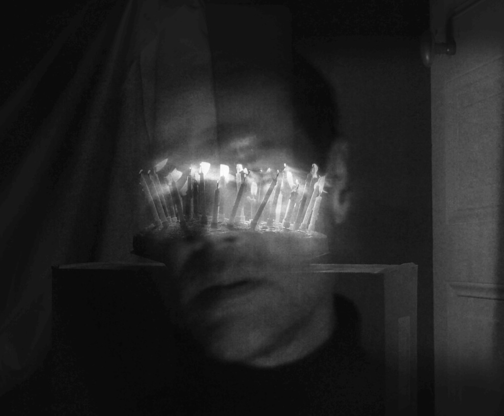 Still from La dernière lueur du soir. A face dissolves behind a row of burning candles in long exposure, black and white.