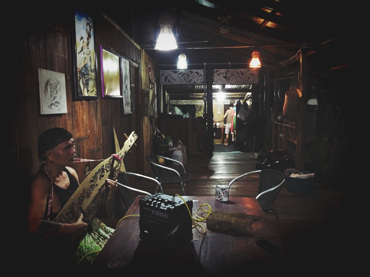 Interior of a traditional longhouse at night. A man sits at the far left playing a sape plugged into a small Roland Cube amplifier. Artworks hang on the wooden walls. A corridor leads to a lit room at the far end.