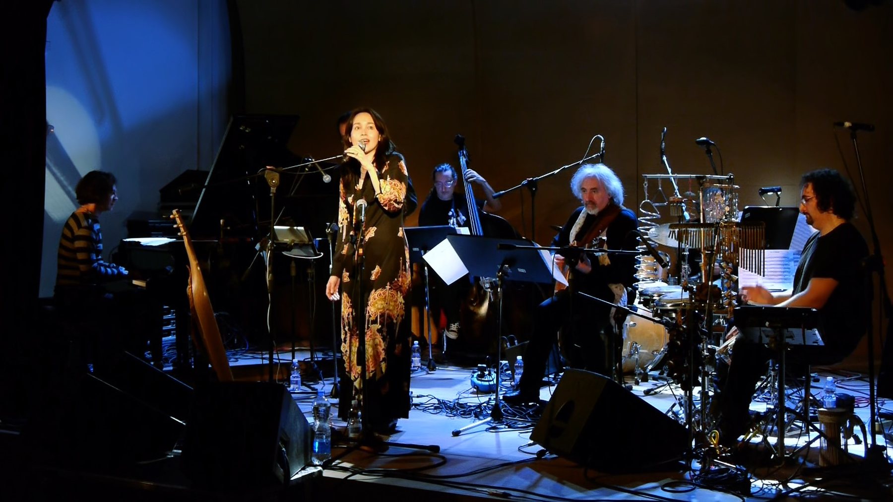 Six musicians on a small stage, piano, upright bass, guitar, and percussion visible, the singer standing centre stage at a microphone.