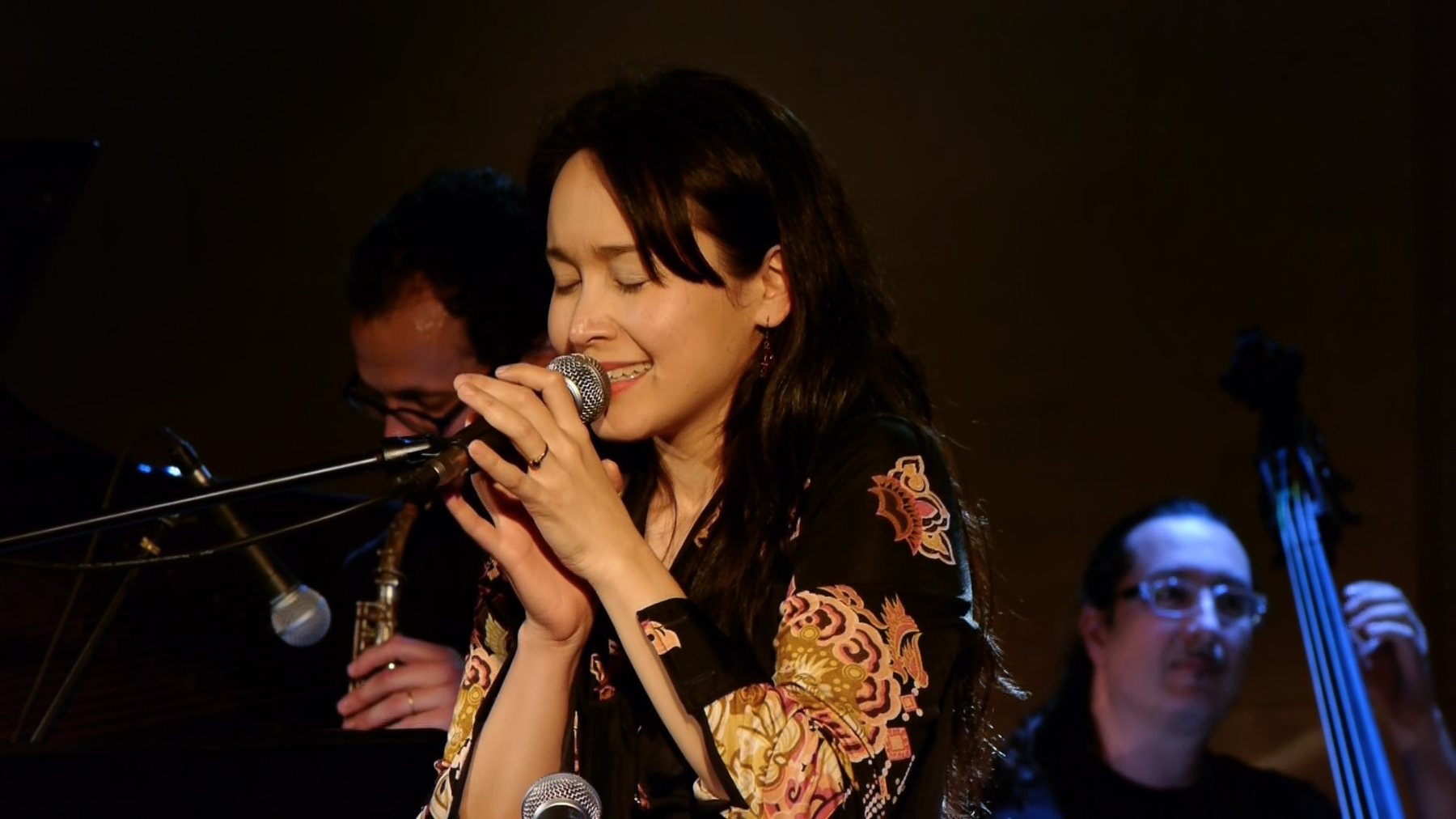 Close-up of a woman singing into a microphone, eyes closed, saxophonist and bassist visible blurred behind her.
