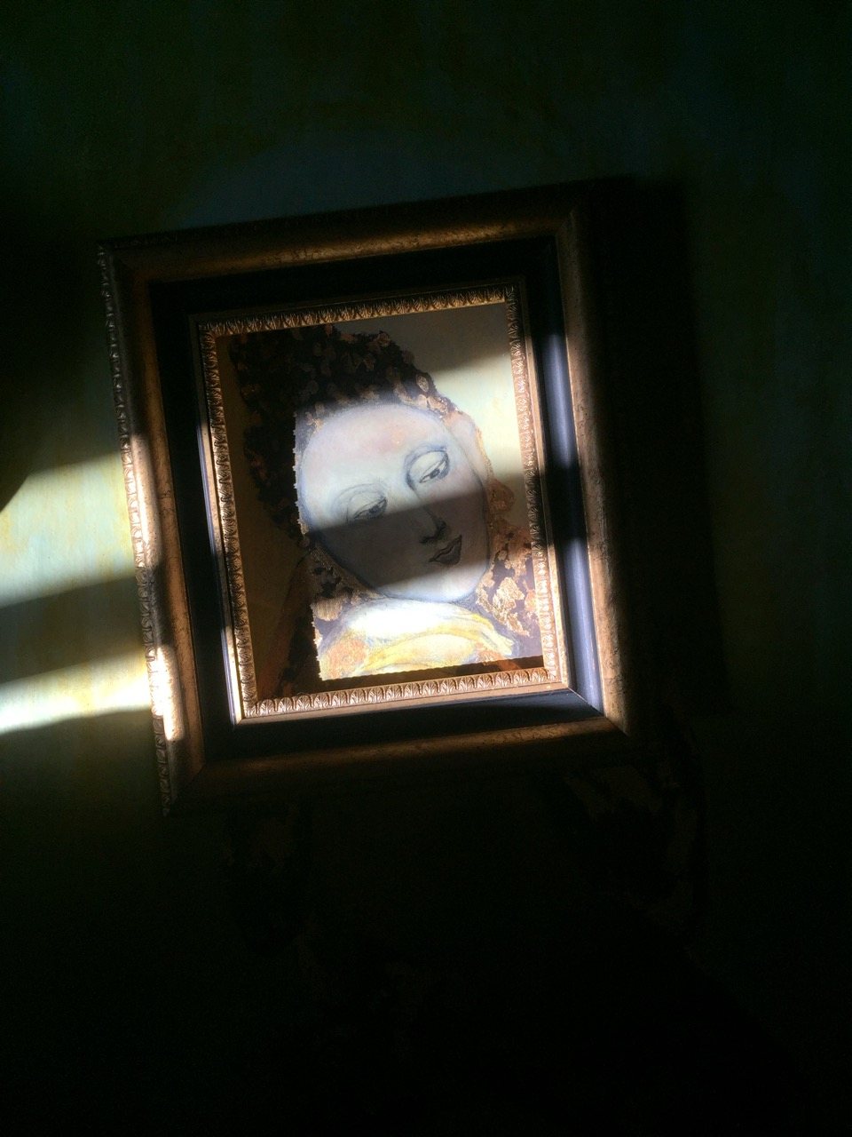 A framed painting of a female sacred figure, eyes closed, gold leaf crown, the face emerging from shadow in late afternoon light.