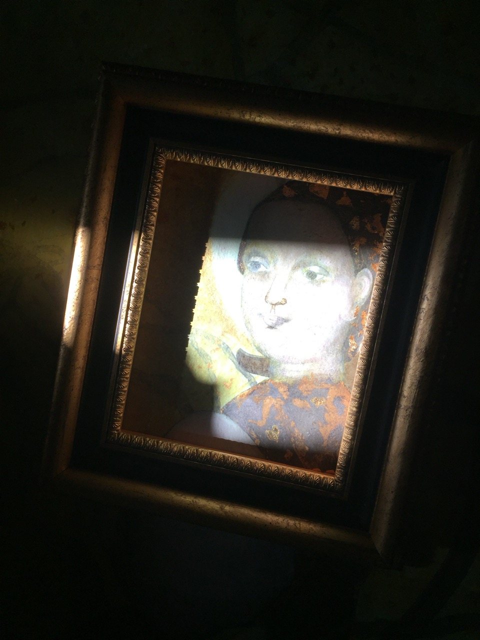 A framed painting of a male sacred figure, pale face, blue garment with gold leaf patterning, caught in a beam of light against a dark background.
