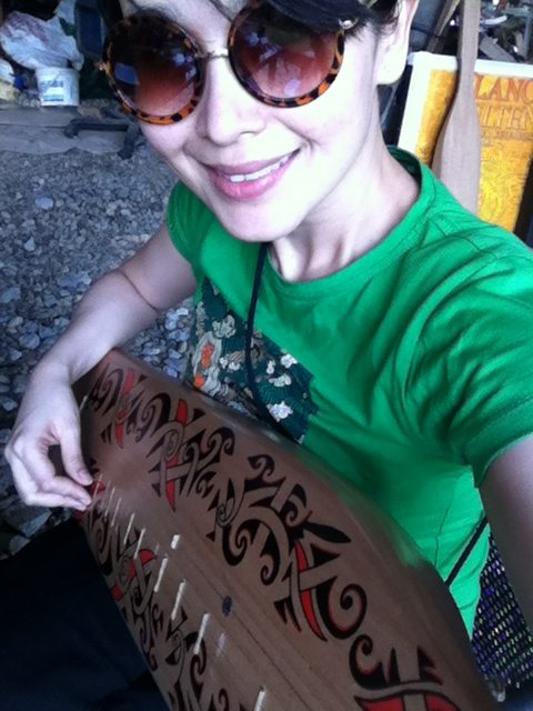 Jacelyn wearing sunglasses and a green shirt, smiling, holding a decorated sape close to the camera so its carved red and black body fills the foreground.