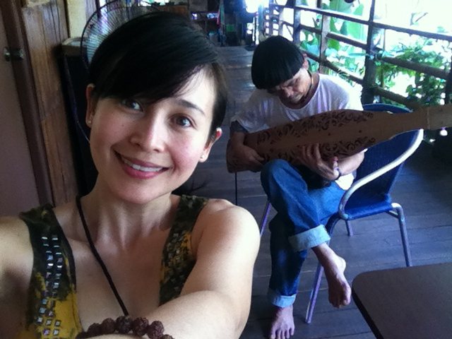 Jacelyn in the foreground smiling at the camera, with Mathew Ngau Jau visible in the background seated and playing his sape on the longhouse veranda.