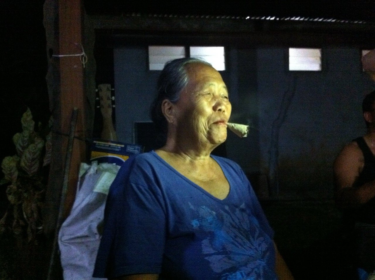 An elderly woman in a blue shirt smoking a cheroot at the evening celebration, her face lit warmly from one side, calm and composed.