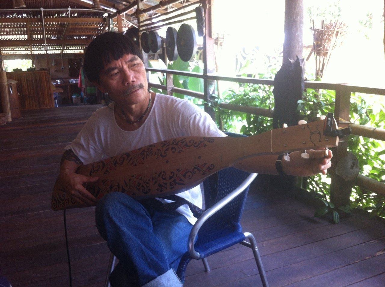 Mathew Ngau Jau seated on the longhouse veranda playing a decorated sape, bronze gongs hanging behind him, the rainforest visible through the railing beyond.