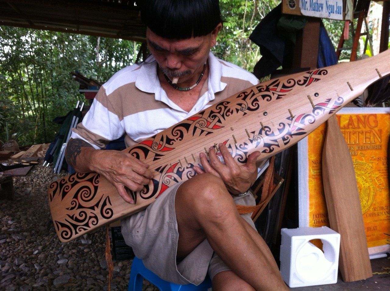Mathew Ngau Jau leaning closely over a newly completed decorated sape, examining it carefully, a sign bearing his name visible in the background.