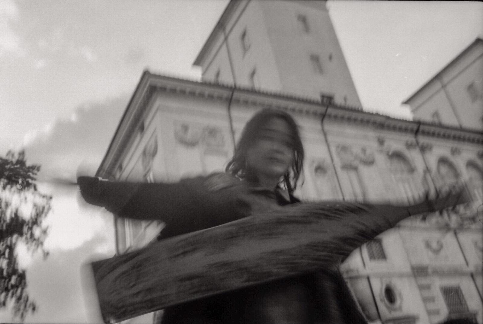 Black and white motion-blur photograph of Jacelyn dancing with the sape raised above her, the facade of the Galleria Borghese behind, the image blurred by movement.