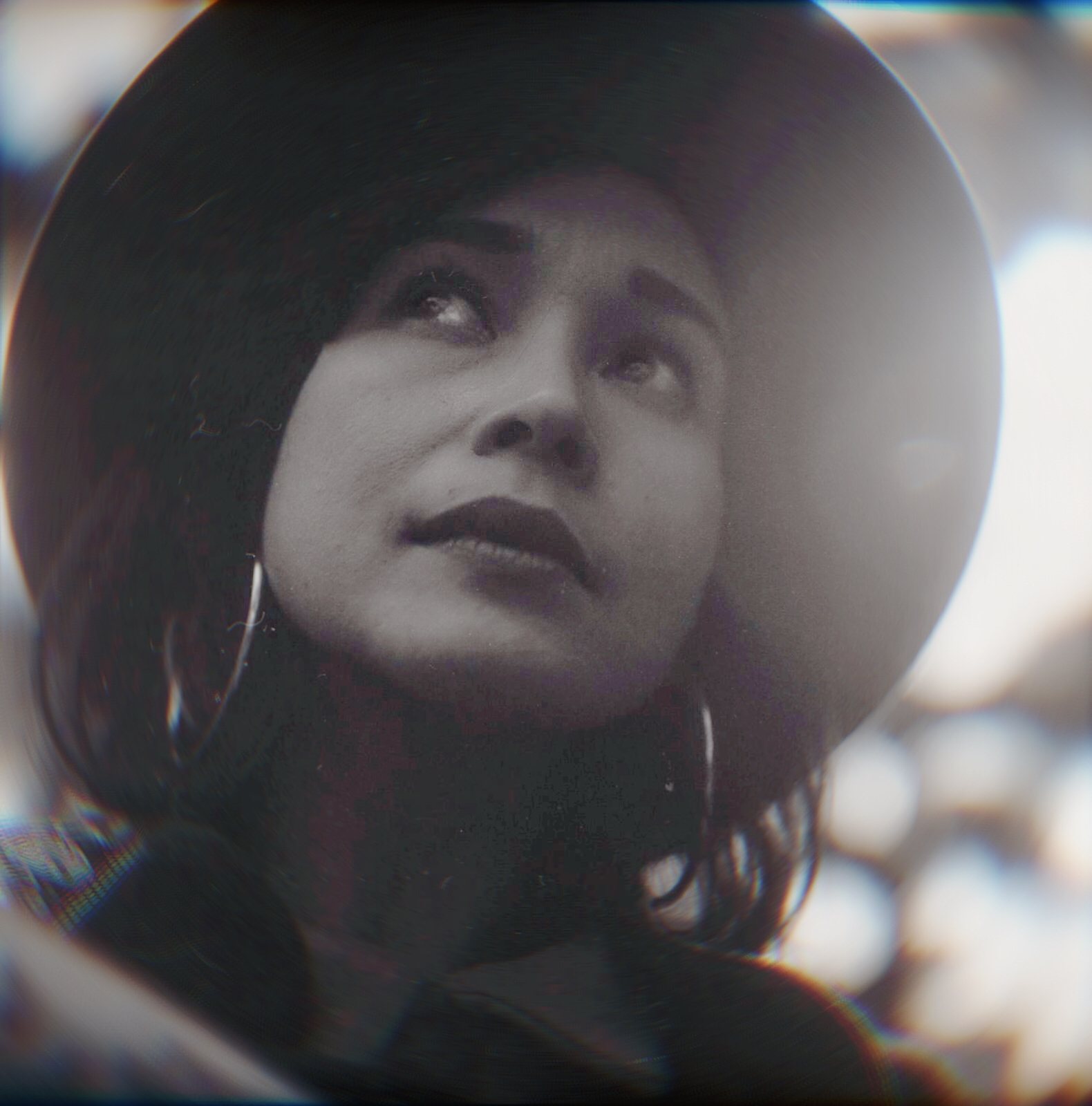 Black and white portrait of Jacelyn looking upward, wide-brimmed hat, hoop earring, bokeh background, taken on film with a Rolleiflex.