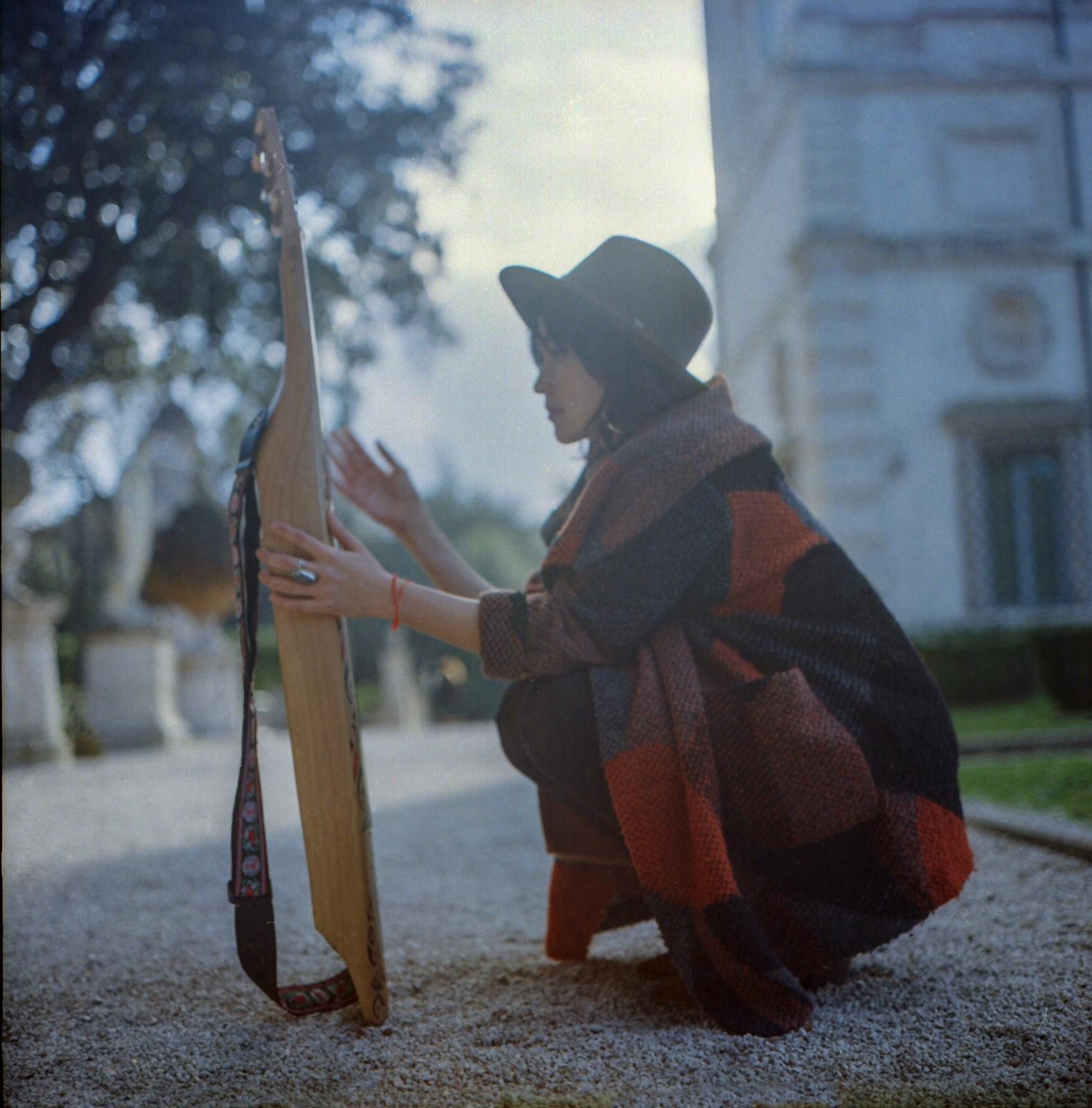 Jacelyn crouching on a gravel path in Villa Borghese, holding the sape upright beside her, the classical facade of the Galleria Borghese visible in the background, winter trees and overcast sky.