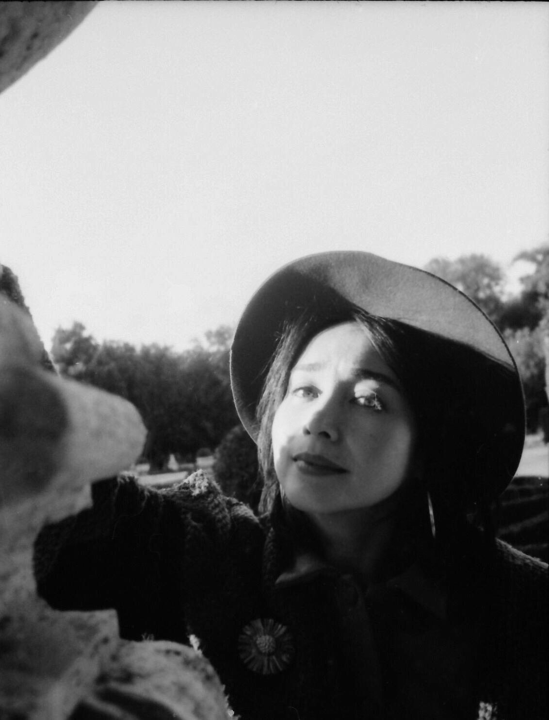 Black and white photograph of Jacelyn with wide-brimmed hat, looking sideways, the neck of the sape visible at lower left, garden and sky behind.