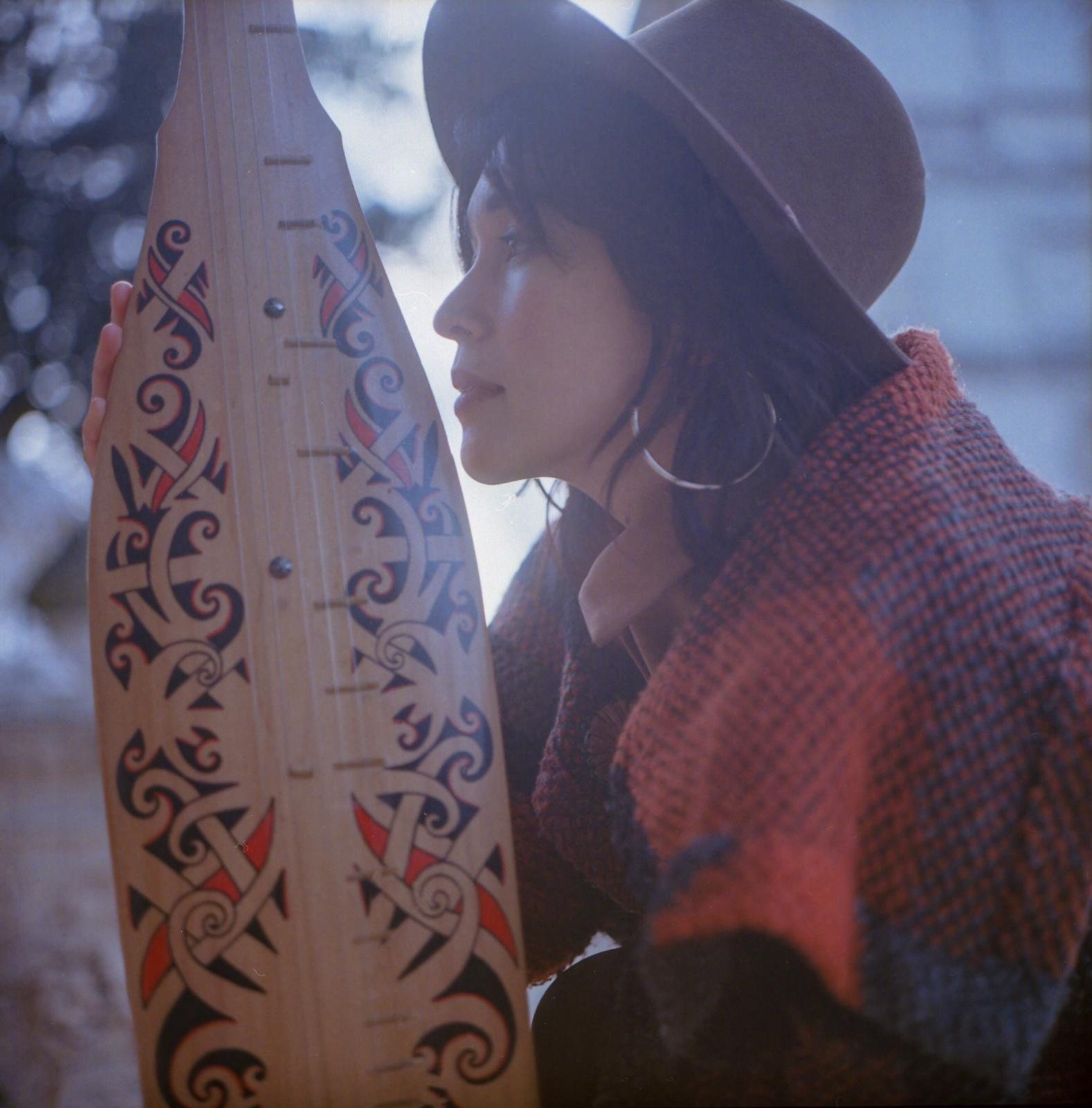 Colour film photograph of Jacelyn in profile, holding the decorated sape body upright close to her face, warm analogue tones, Villa Borghese garden behind.