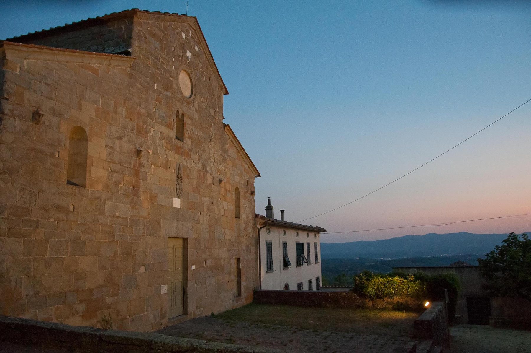 The exterior of the stone church at dusk, a warm light beside the entrance, hills in the distance.