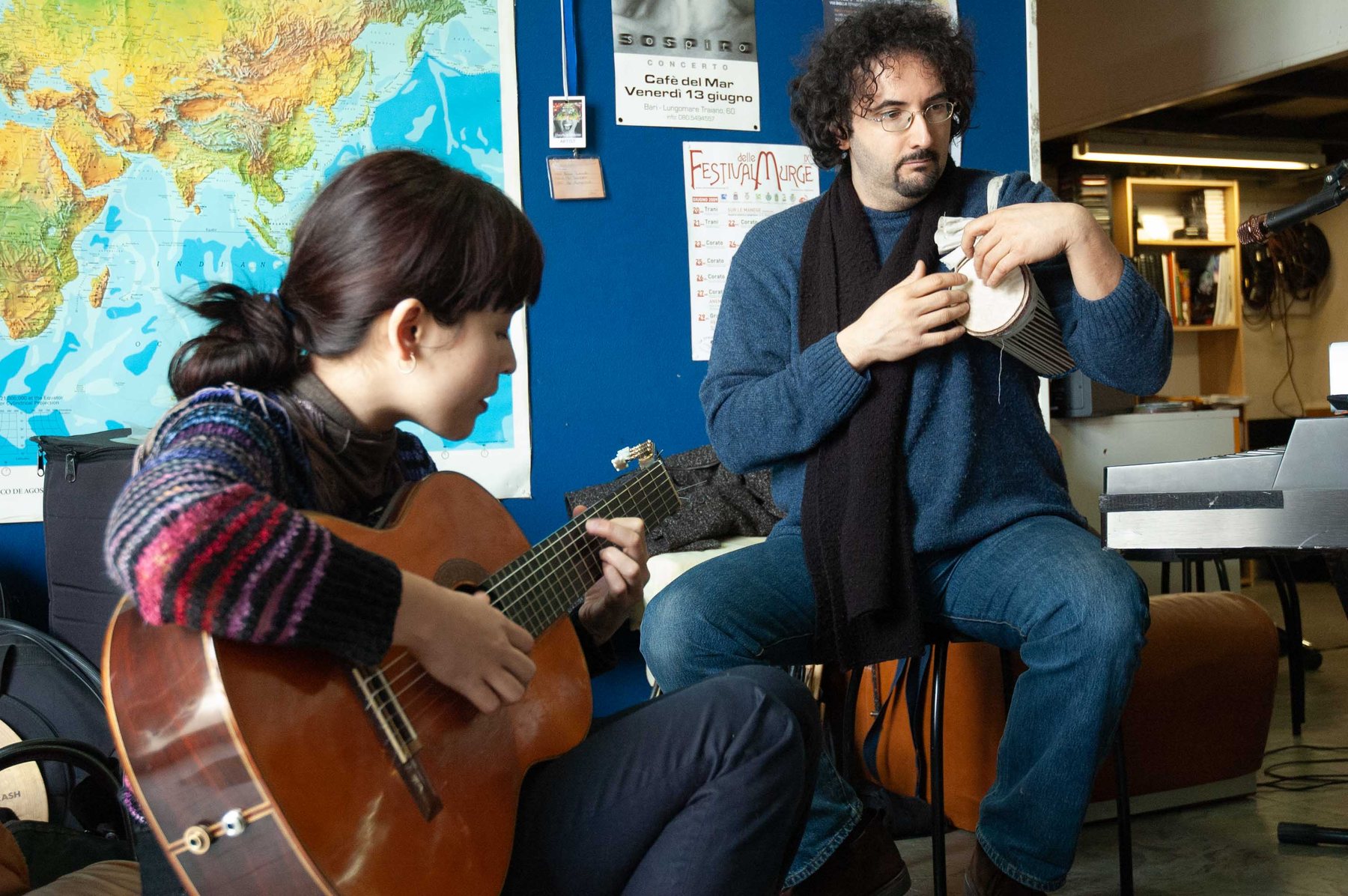 Jacelyn playing guitar with a percussionist beside her, a world map and concert posters on the blue wall behind them.