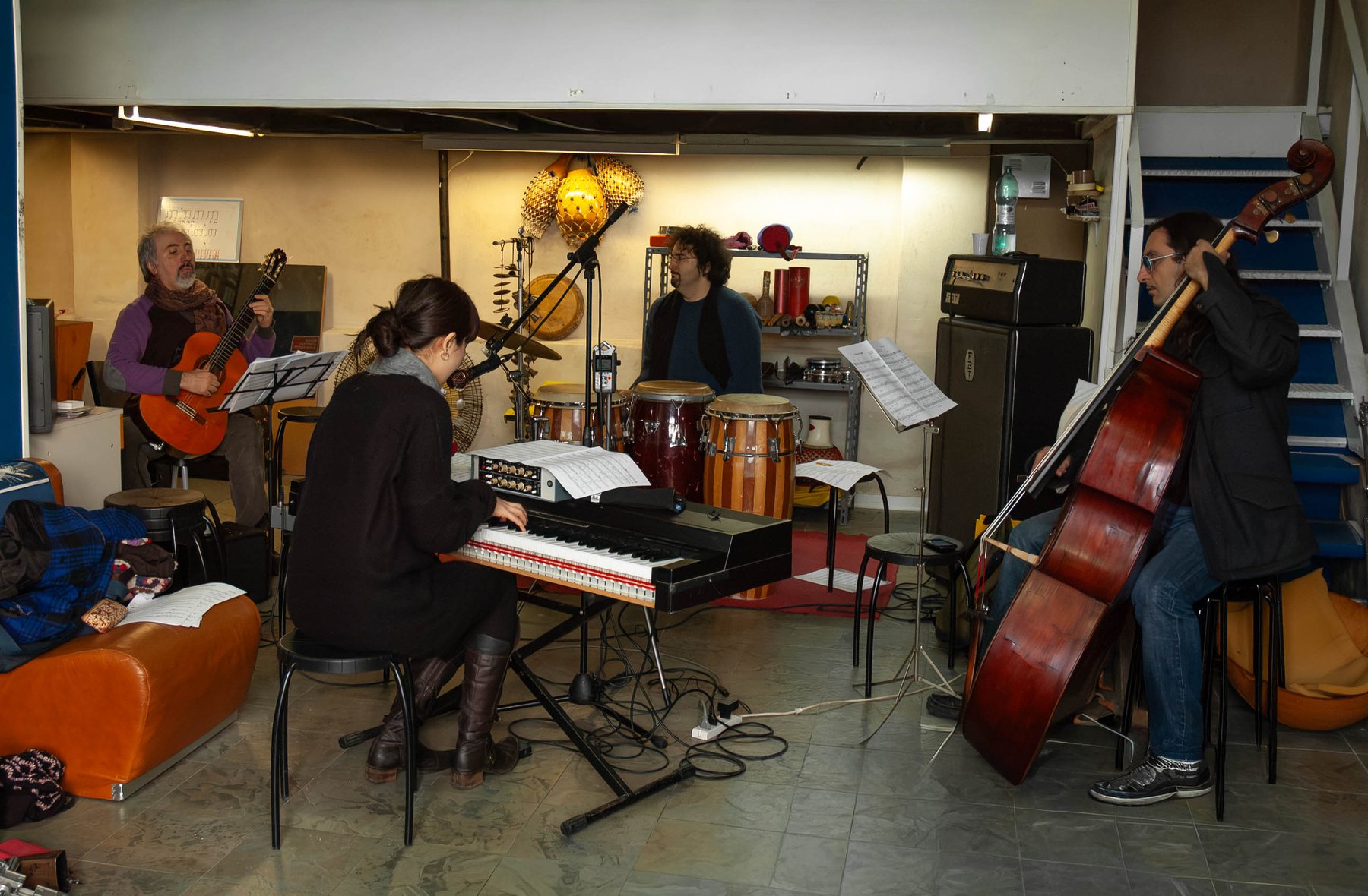 Full ensemble rehearsal in a Puglia studio: Jacelyn at a keyboard, guitarist reading a score, percussionist behind a conga drum, double bassist to the right, music stands and cables throughout the room.