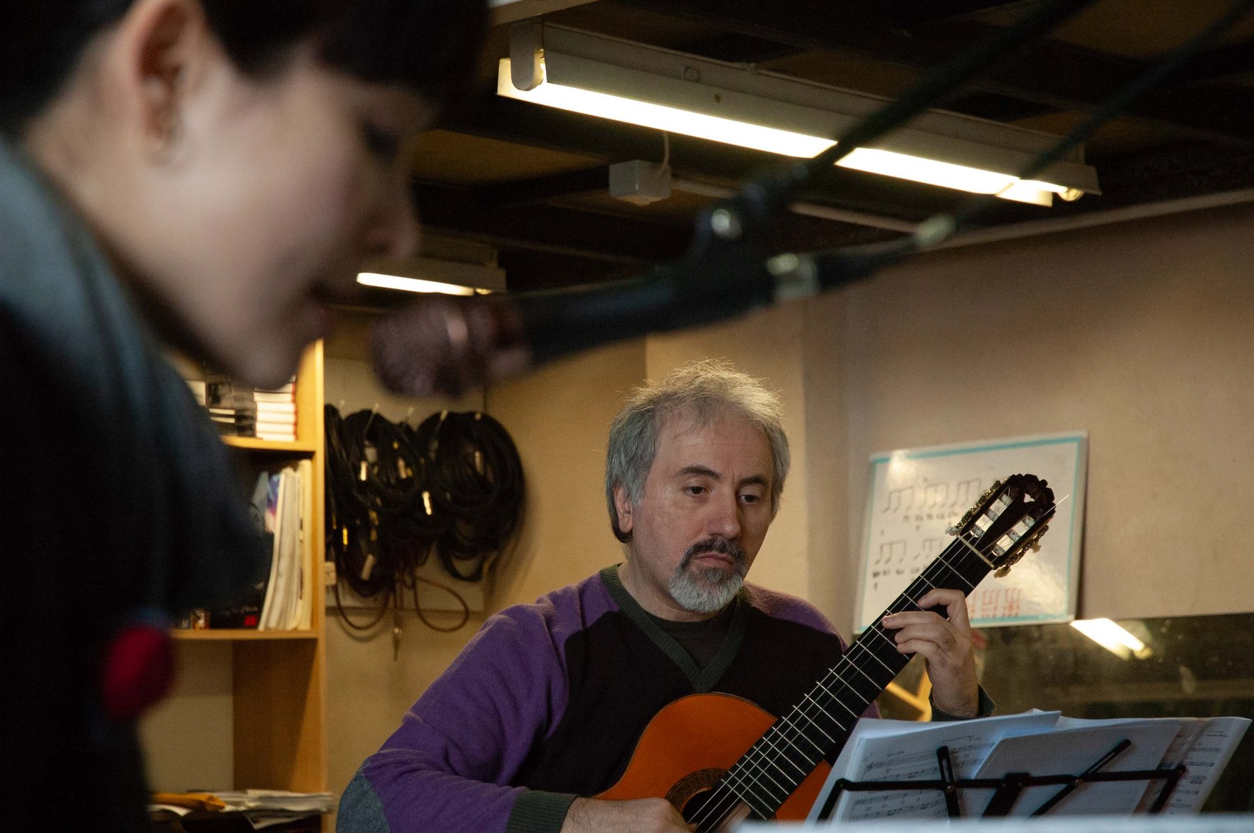 A guitarist studying a score on a music stand in a rehearsal room, concentrating, a blurred figure in the foreground.