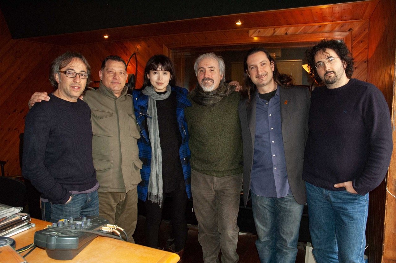 All six Fortressa musicians standing together in the warm wood-panelled studio after a session, looking at the camera, relaxed and composed.