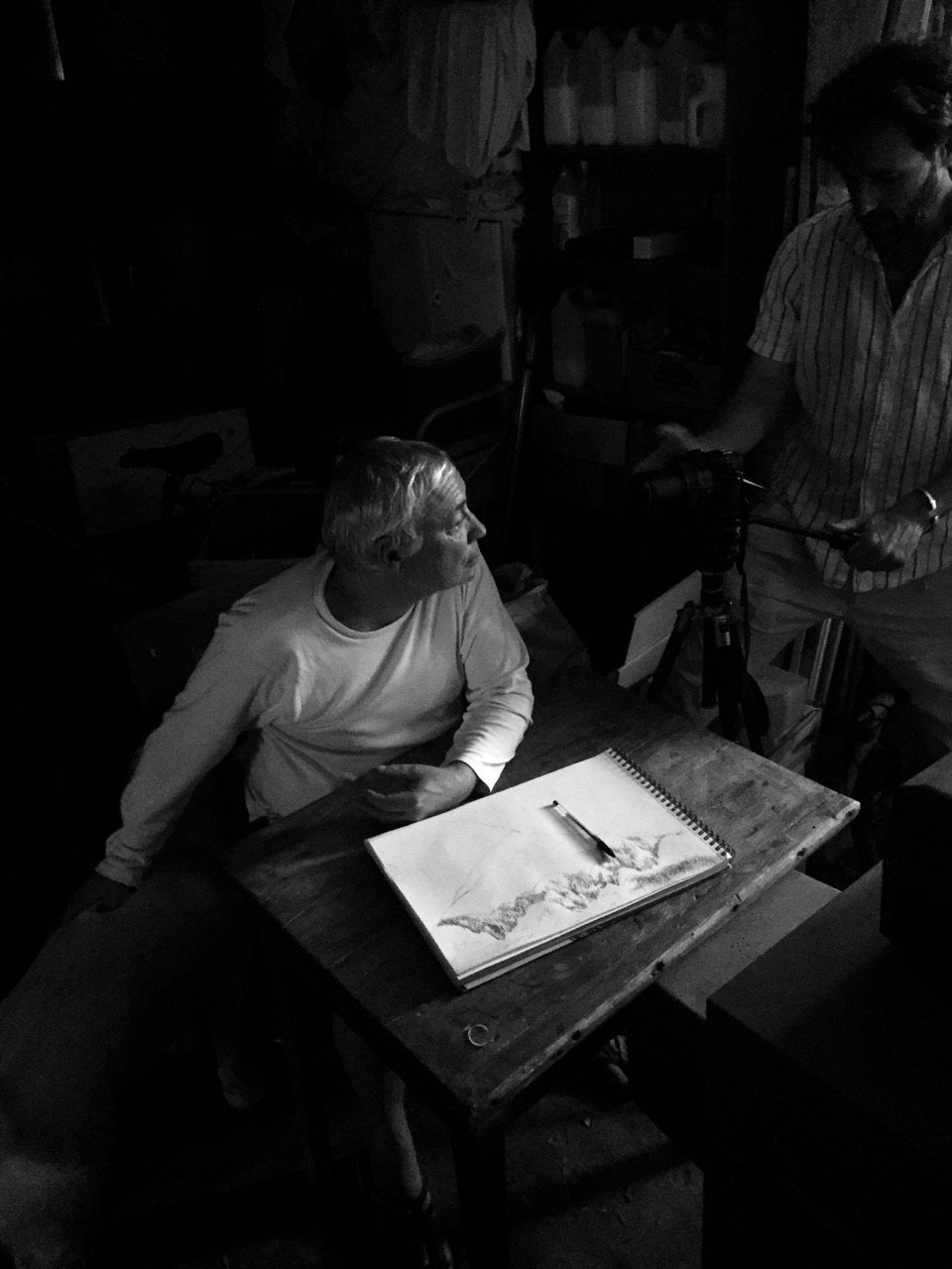 The artist seated at a workbench in low light, sketching a mountain range in a spiral notebook, a crew member visible behind him.