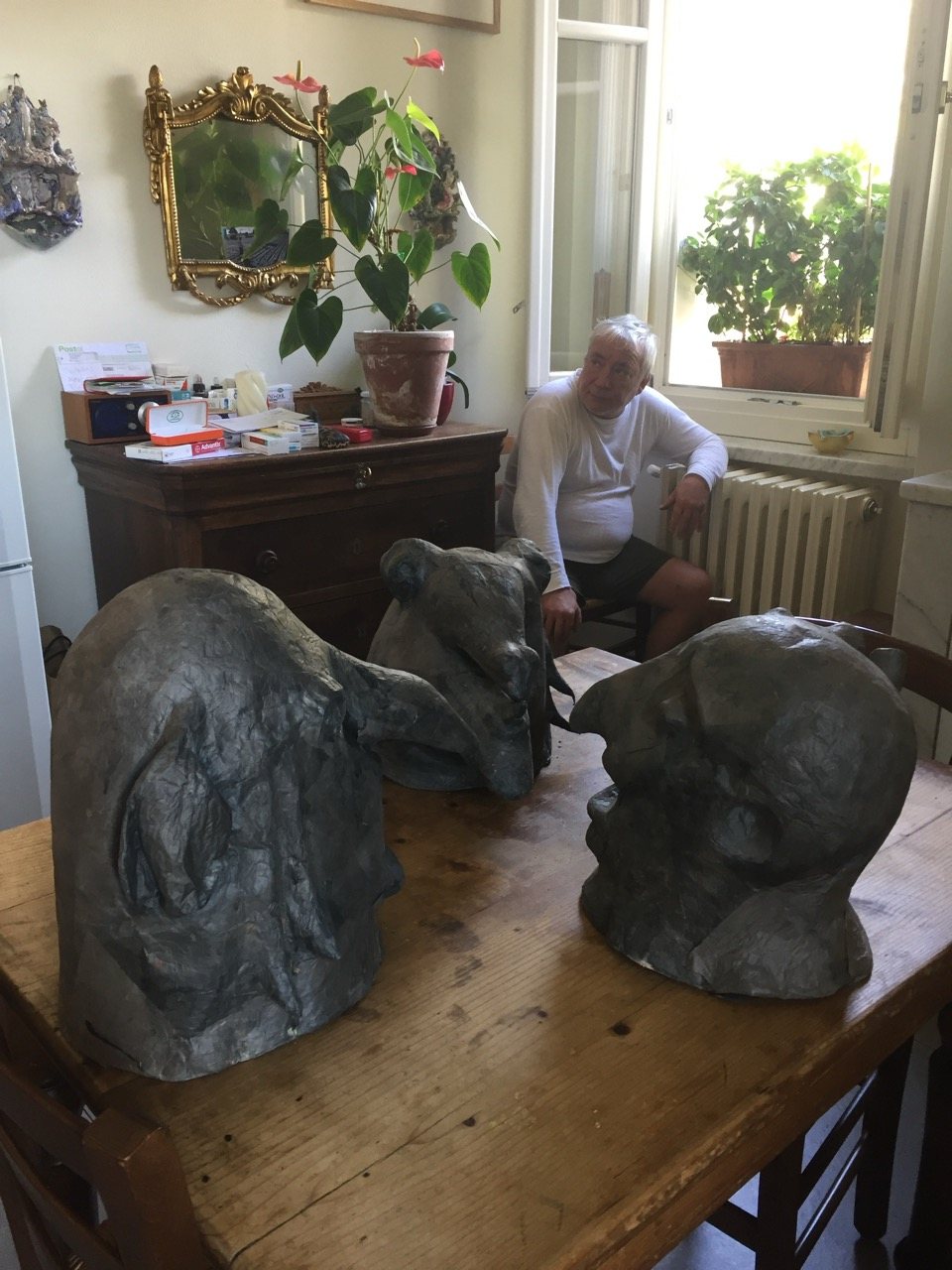The artist seated in his studio, three sculpted heads arranged on the table before him, window light behind.