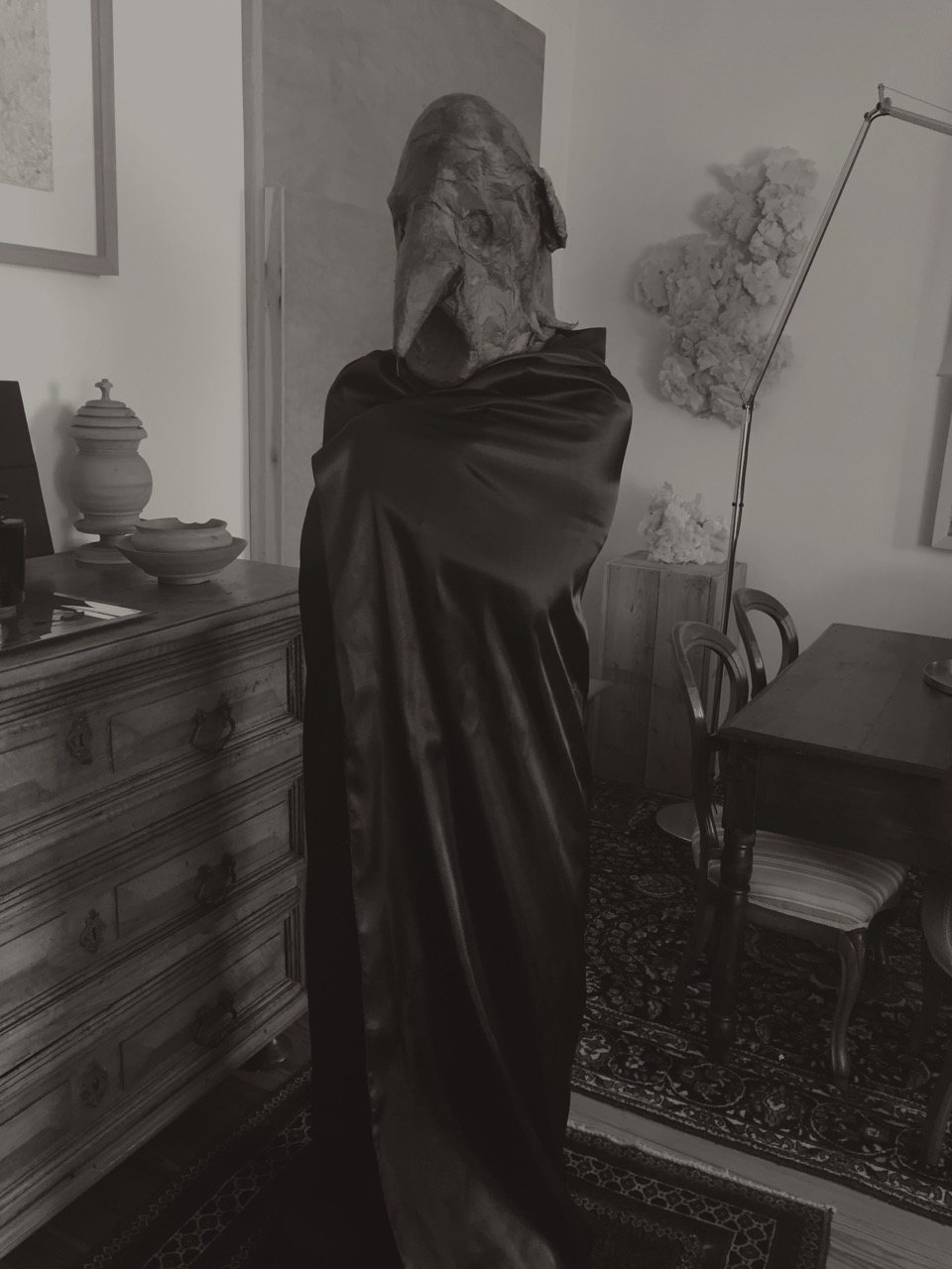 A cloaked, bird-masked figure standing in a quiet domestic interior, an antique chest of drawers and soft furnishings visible around it.