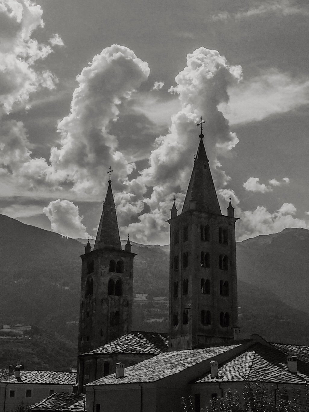 Two Romanesque bell towers against a sky filled with dramatic clouds, mountains visible in the distance.