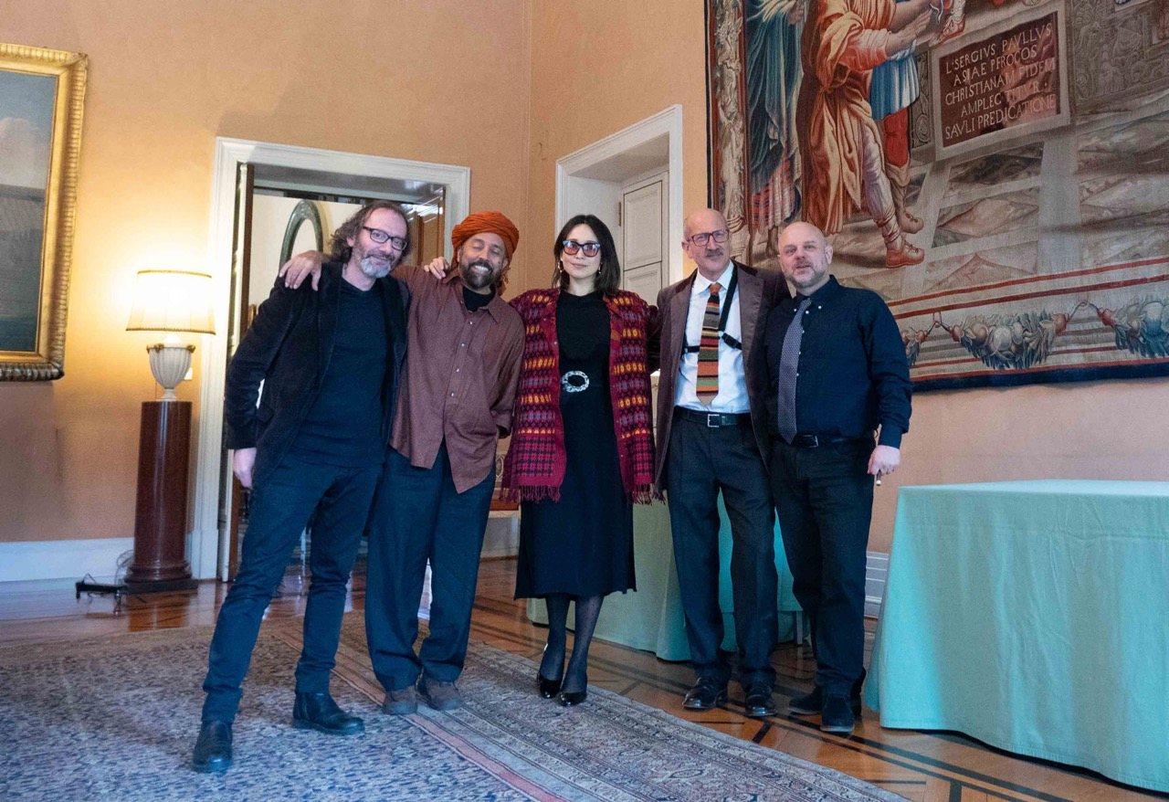 Five musicians standing together and smiling in a formal reception room, tapestries and a gilt-framed painting on the wall behind them.