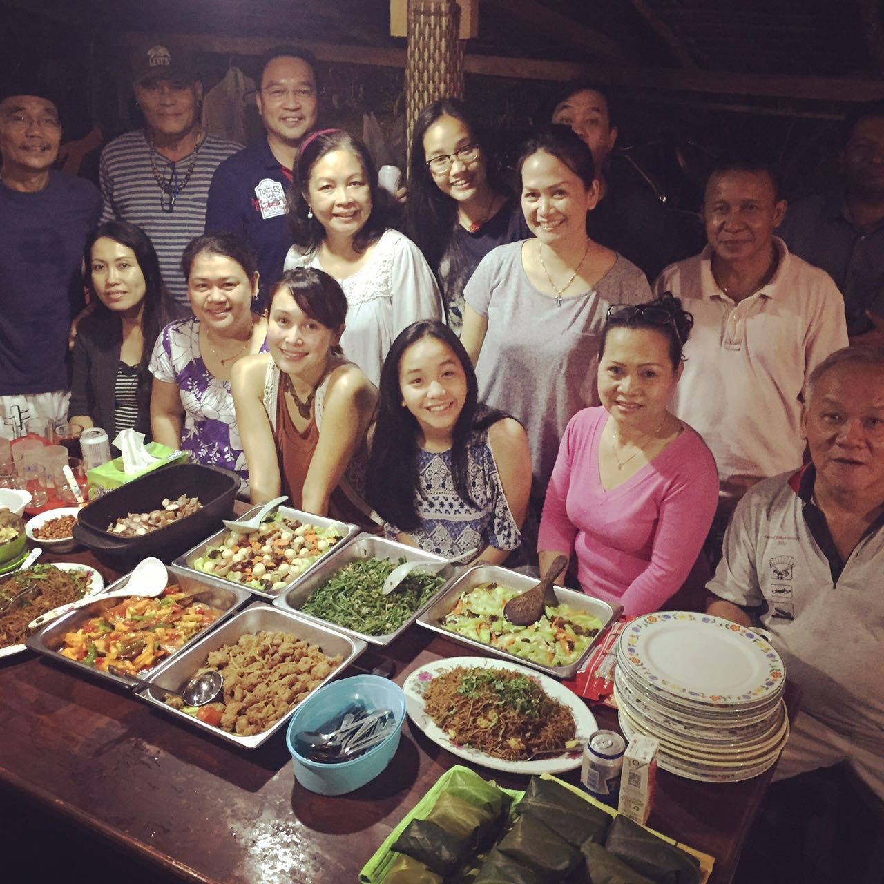 A large group of people gathered around a long table covered with food, smiling at the camera inside a wooden longhouse at night.