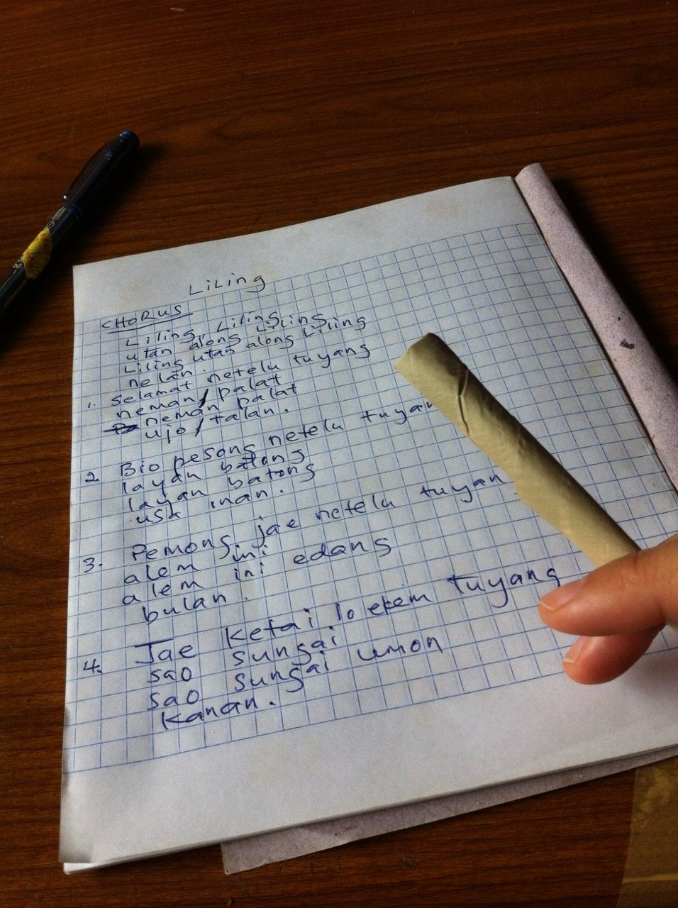 A handwritten page of lyrics to Leling on squared notebook paper, in blue ink, with the chorus and four numbered verses, a pen resting beside the paper on a wooden table.