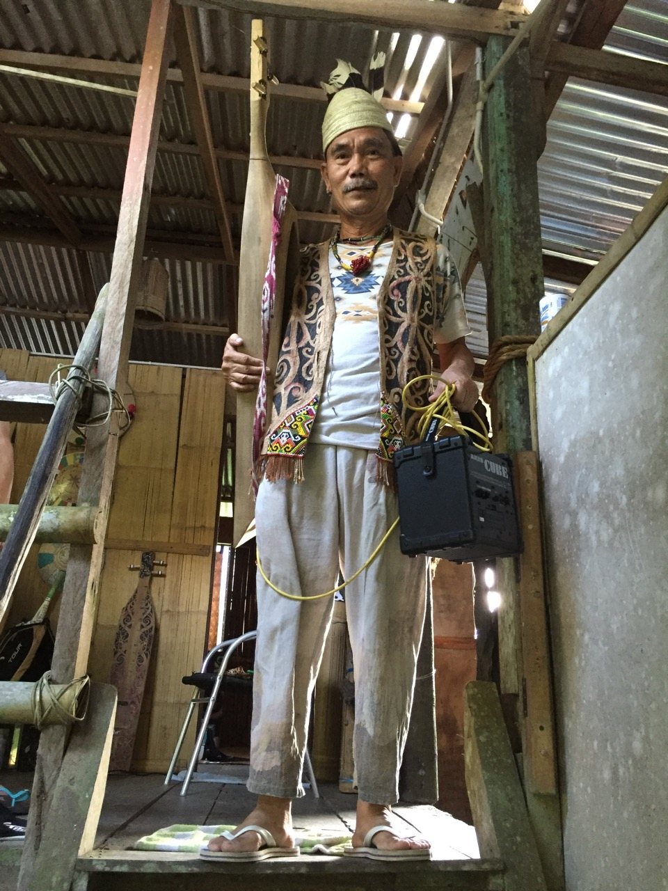 Mathew standing on a wooden staircase inside a barn-like space, wearing traditional dress, holding a sape in one hand and a Roland Cube amplifier in the other, cables draped over his shoulder.