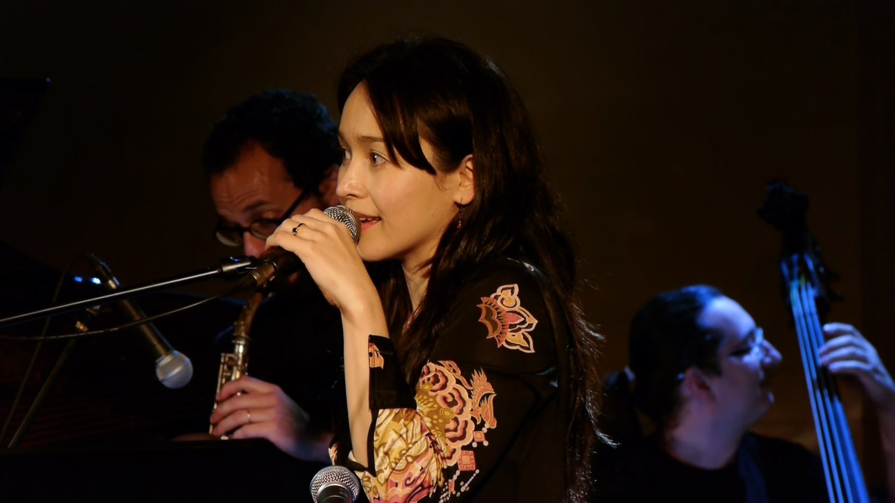 A woman singing in profile at a microphone, a saxophonist and bassist visible beside her in the low stage light.