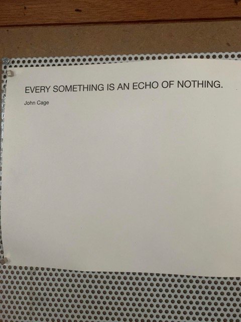 A printed page pinned to a pegboard wall in the studio, reading: Every something is an echo of nothing, John Cage.