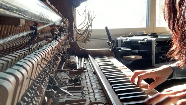 Hands playing the prepared piano, exposed hammers and strings in the foreground, window light behind, recording equipment beside the keys.