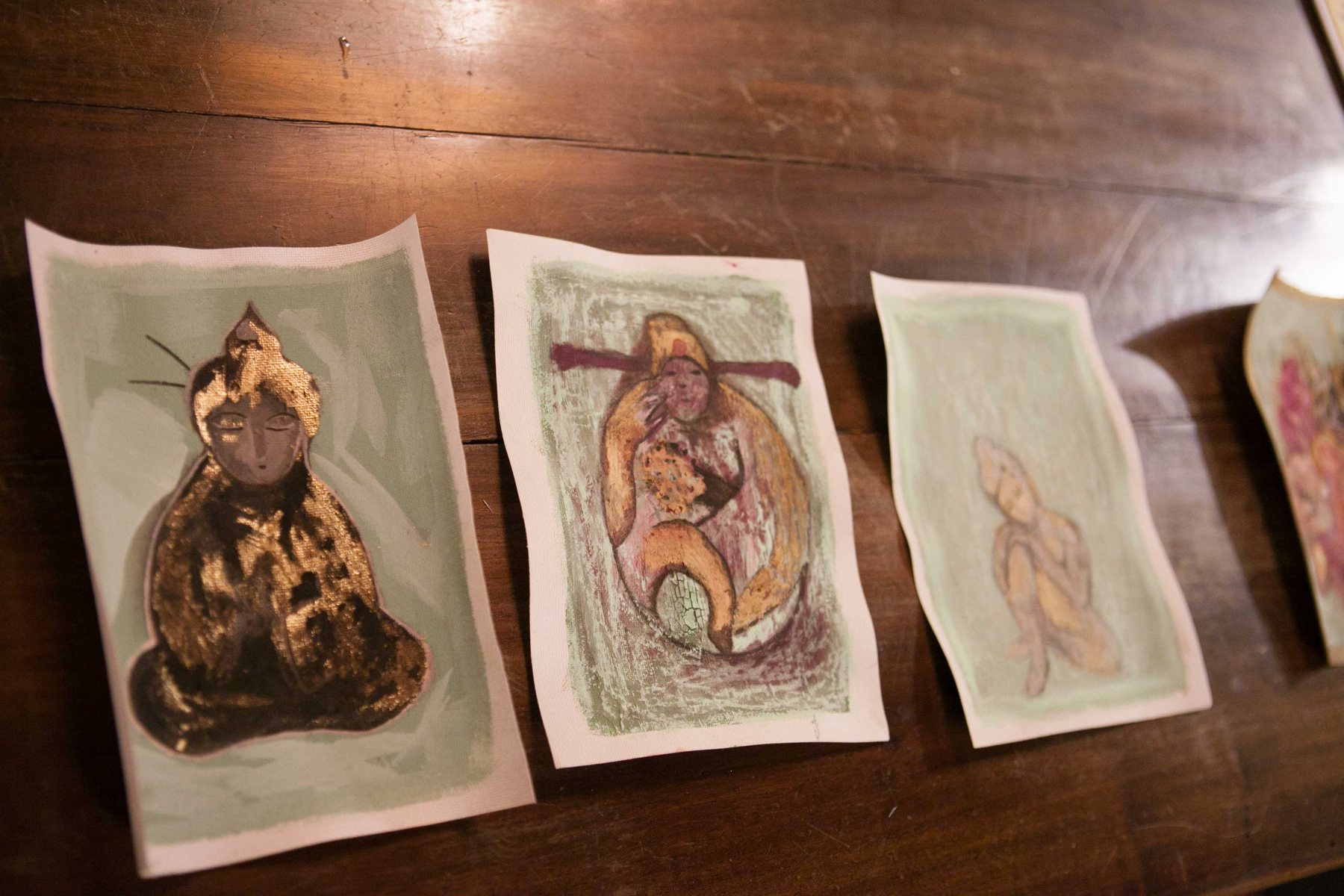 Three small painted figures on paper, laid out on a wooden table, one in gold leaf, one holding a small form, one unfinished.