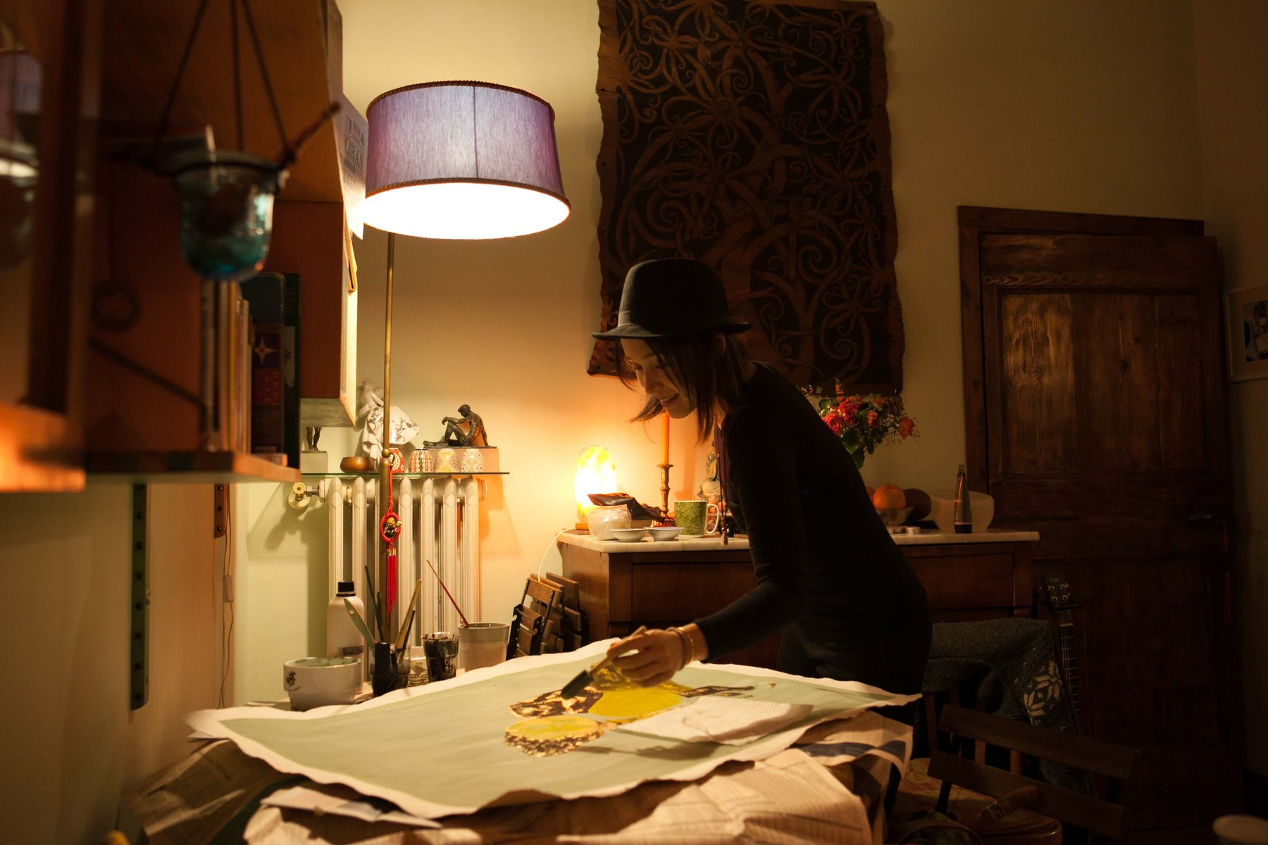 A woman leaning over a painting on a table, lit by a warm lamp, absorbed in the work.