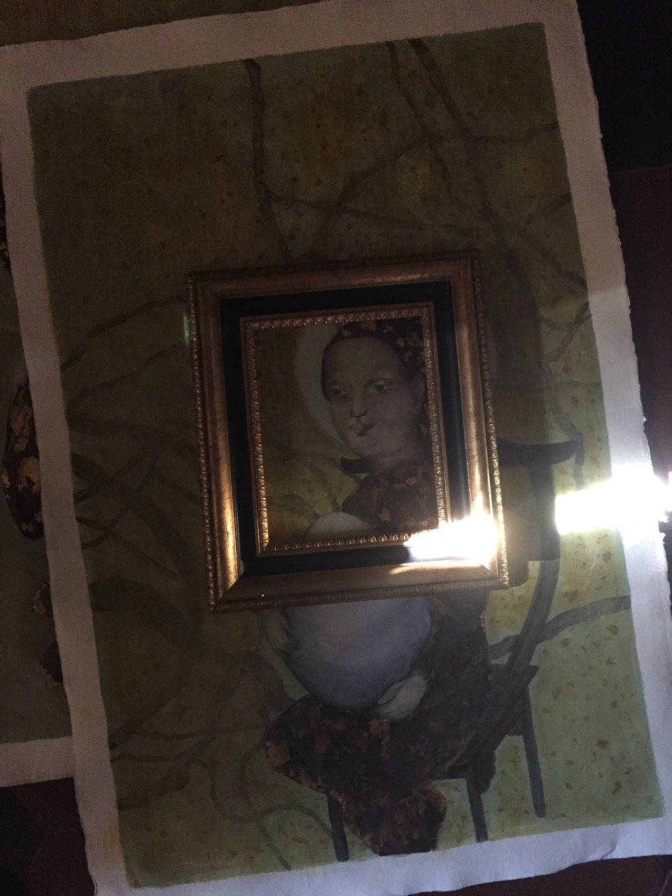 A small framed painting of a female figure hung against patterned wallpaper, lit from below by candlelight, the frame glowing at its lower edge.