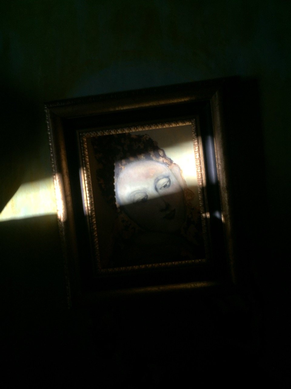 A framed painting in near-darkness, a face just barely visible beneath gold leaf and shadow, caught by a narrow beam of light.