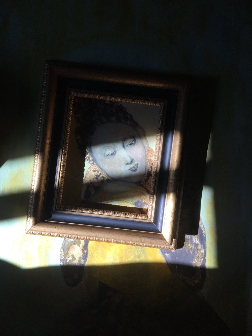 The same framed sacred figure painting in fuller light, gold and blue visible, the face now fully present, the frame casting a long shadow.