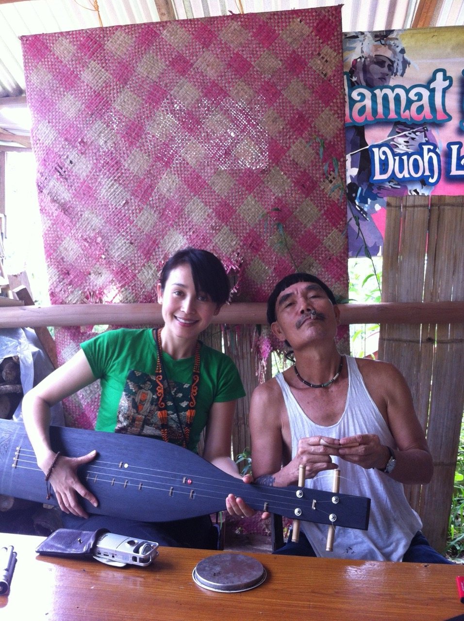 Jacelyn and Mathew Ngau Jau sitting side by side, both holding a sape together and looking at the camera, a colourful woven mat and a welcome banner visible on the wall behind them.