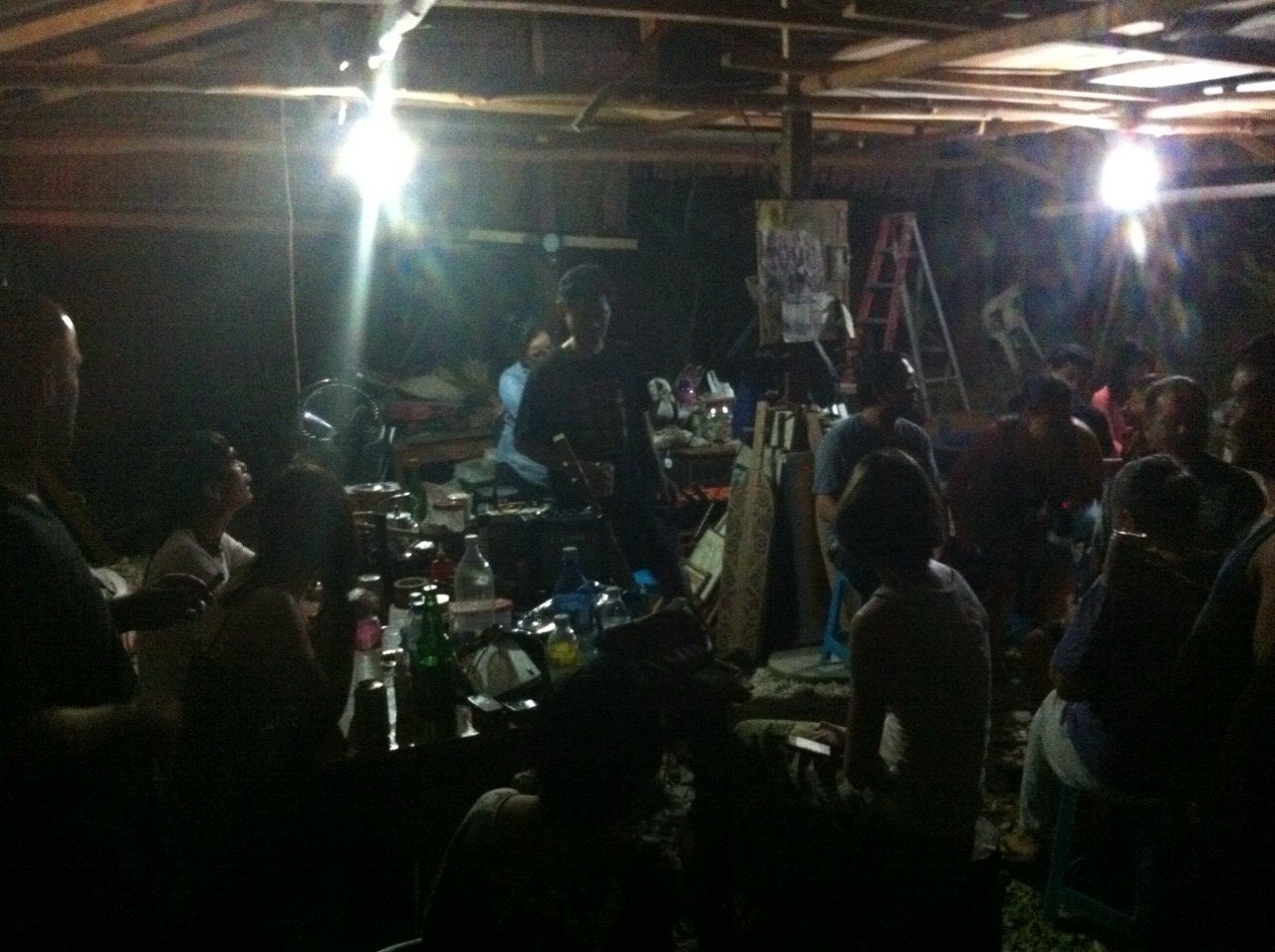 The interior of the longhouse at night, lit by two bright overhead lights, a crowd of people gathered watching a performance, drinks on tables, an atmosphere of celebration.