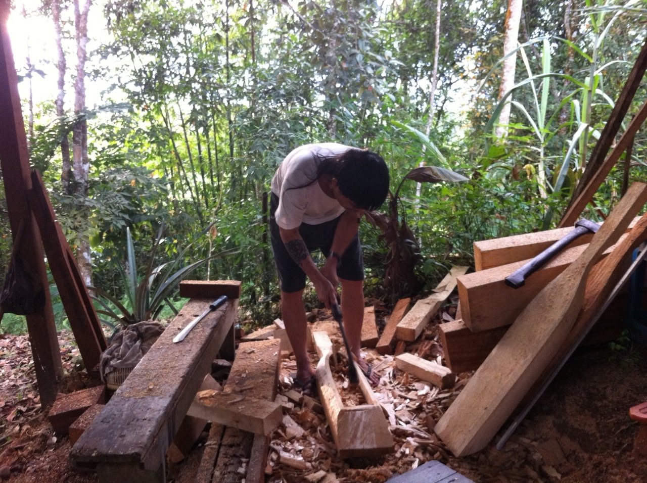 Mathew Ngau Jau bent low over a long piece of timber in an open forest clearing, surrounded by wood shavings and planks, shaping the body of the sape with a hand chisel.