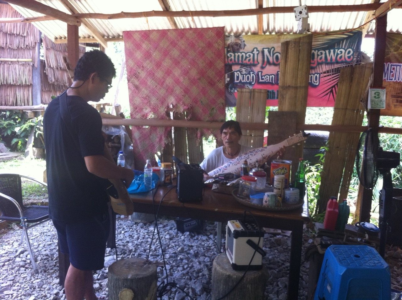 Mathew Ngau Jau seated at his workshop stall with a decorated sape laid out on the table between him and a young man standing opposite, a welcome banner and a pink woven mat visible behind them.